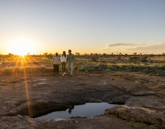 Three people stand near a natural water hole or rock pool on Patji Lands at sunrise. The sun appears on the horizon, creating dramatic sun rays and golden light across the desert landscape. Their silhouettes are framed against the warm sky, with desert vegetation visible in the background.