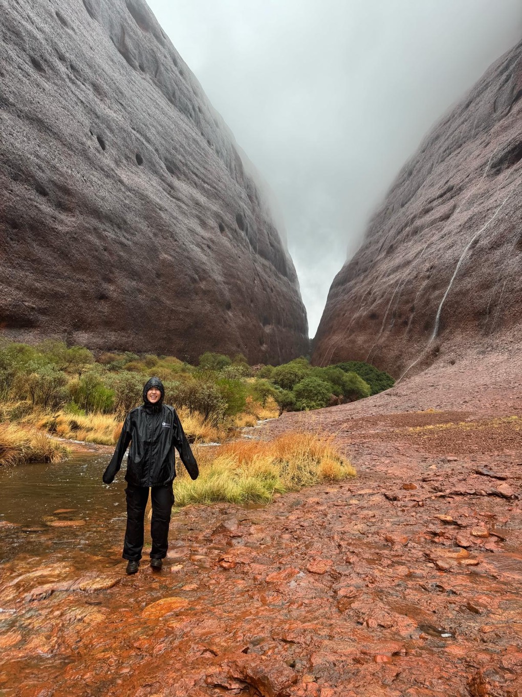 Rachael is wearing waterproof, surrounded by mini waterfalls running down the large rock on either side, with thick clouds in the sky covering the top of the rock, a rare sight – rain at Walpa Gorge.