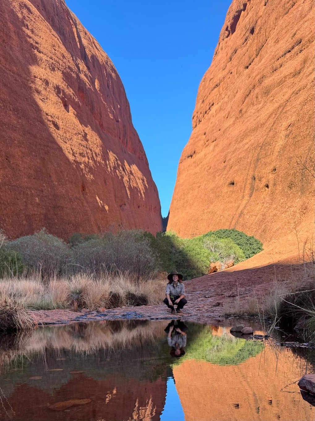 Racheal crouching near a watering hole at Walpa Gorge. On either side are the mighty walls made of red rock contrasting against the bright blue sky.