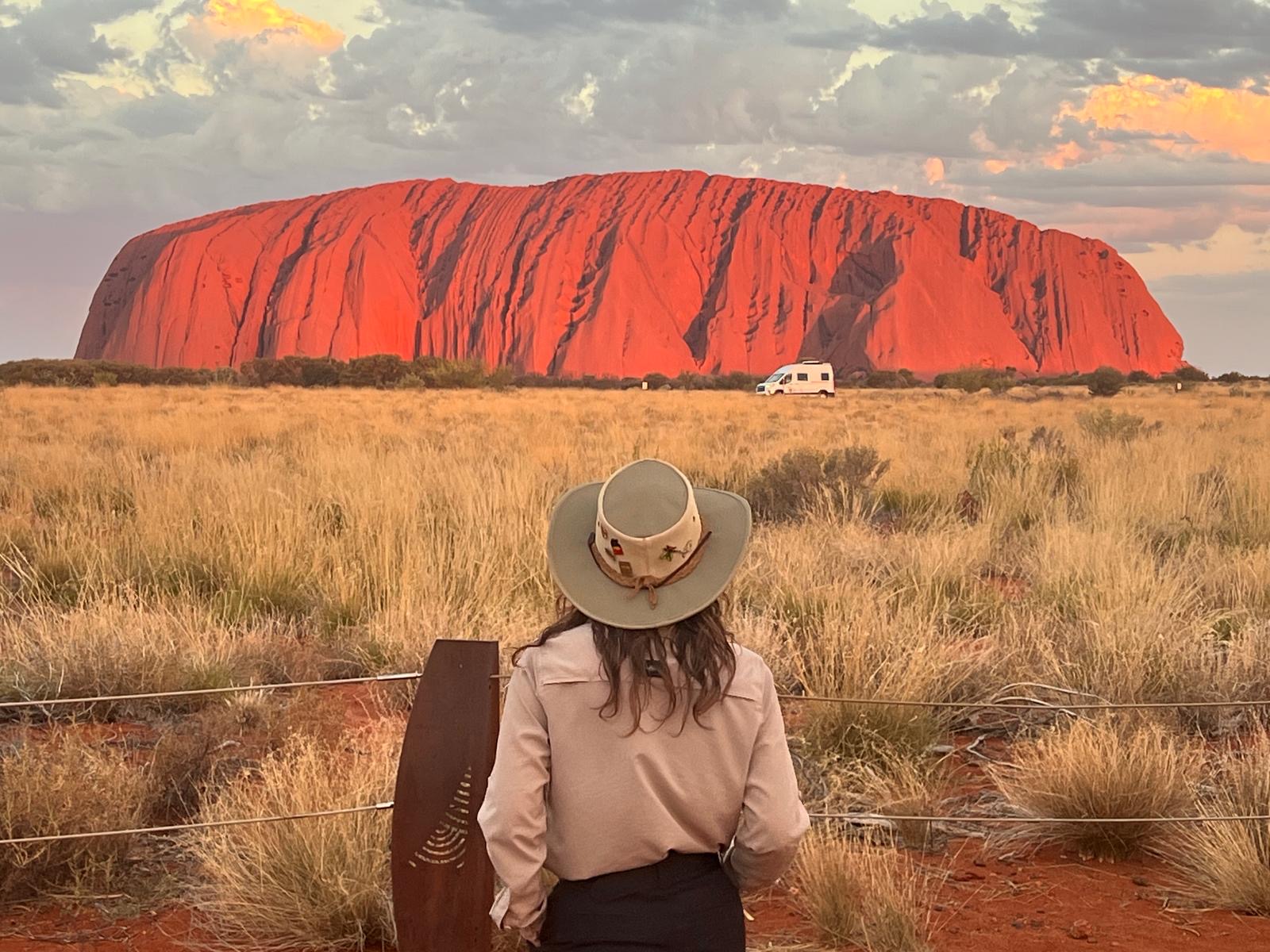 Rachael looking at Uluru, from a distance, with long dried grass in shades of green in between. Uluru is bright red, as the sun shines against it, highlighting large shadows in the rock.