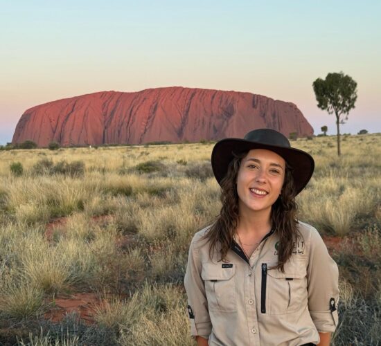 Rachael, SEIT guide, smiling in front of Uluru as the sun sets, showing beautiful pink, yellow, and blue colours in the sky with green grass bushland in the foreground.