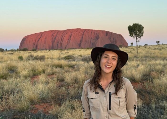 Rachael, SEIT guide, smiling in front of Uluru as the sun sets, showing beautiful pink, yellow, and blue colours in the sky with green grass bushland in the foreground.