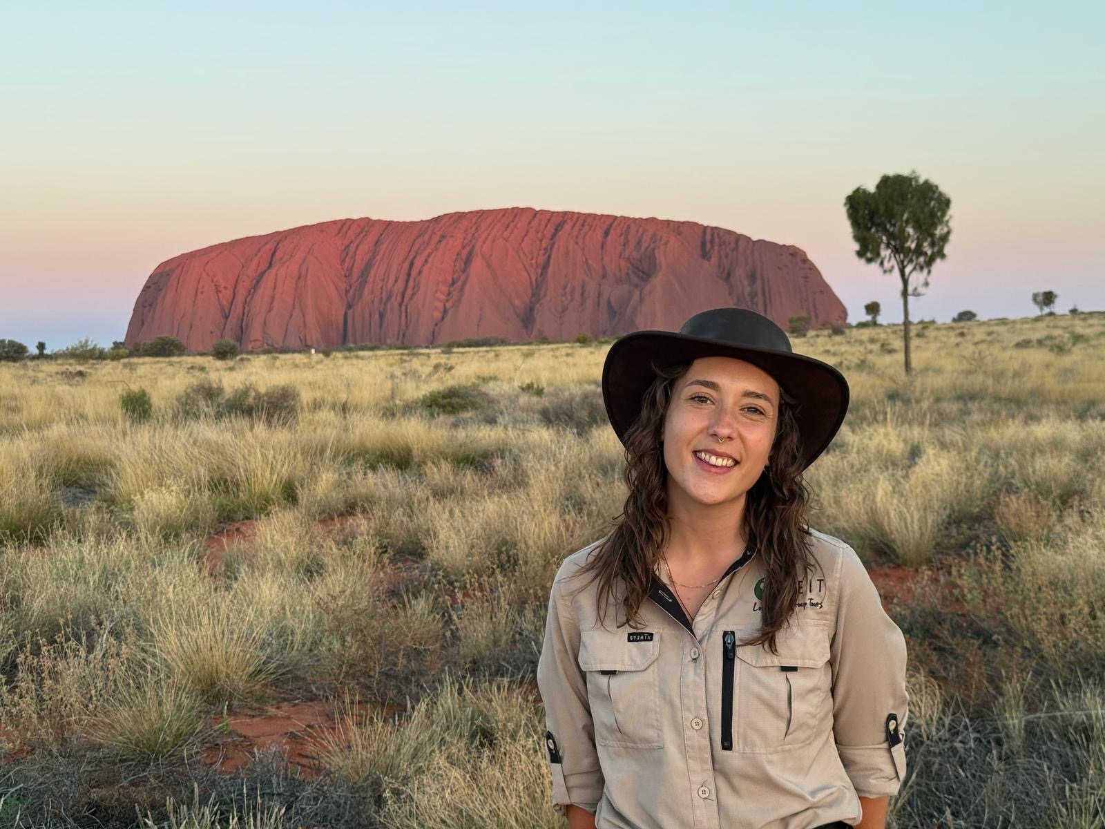 Rachael, SEIT guide, smiling in front of Uluru as the sun sets, showing beautiful pink, yellow, and blue colours in the sky with green grass bushland in the foreground.
