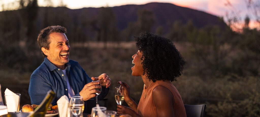Two people laughing over dinner with Uluru in the background